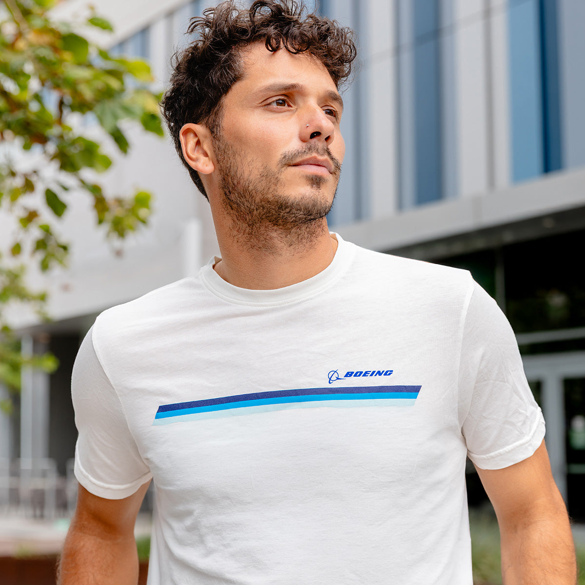 A man with short curly hair and a trimmed beard wears the Boeing Unisex Signature Logo Stripe T-Shirt by BOEING, standing outdoors near a modern glass building with greenery in the background.