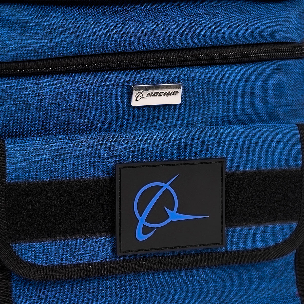 The Boeing Double Deck Lunch Box, from The Boeing Store, is a blue fabric bag featuring a metal Boeing plaque on top and a larger blue logo with a stylized plane on a black patch. It has a black horizontal strip in the middle and comes with a removable shoulder strap for easy carrying.