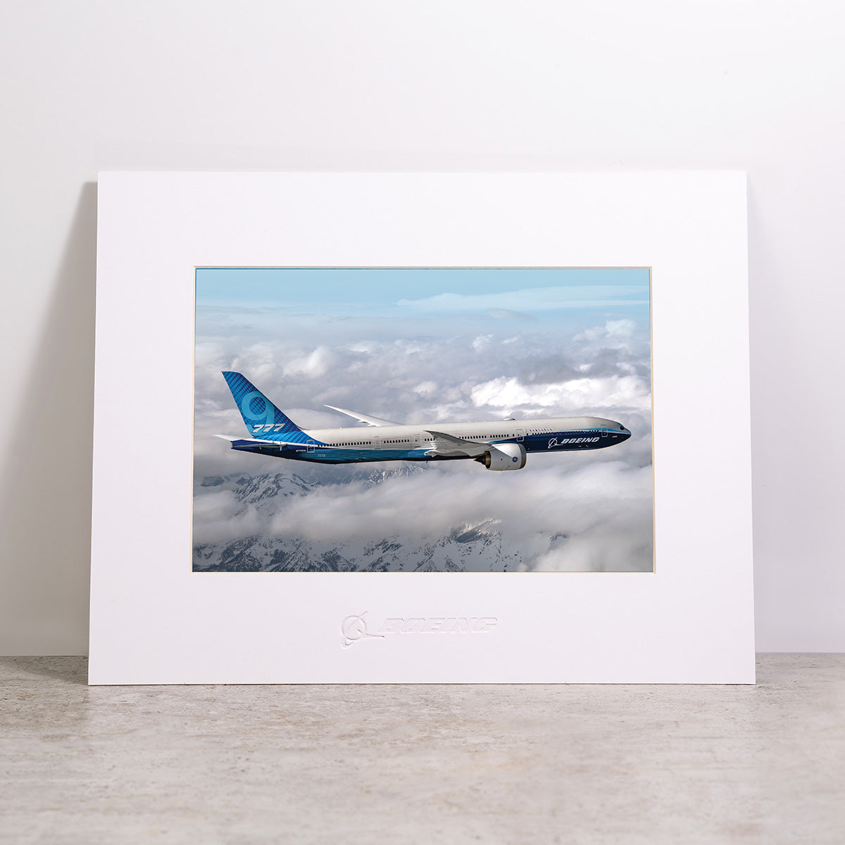 Showcased on a light surface against a plain white background, the Boeing 777-9 Matted Print - Small by Boeing features a high-resolution image of a Boeing 777 airplane soaring above the clouds. This elegant matted print delivers exceptional clarity through digital printing and proudly displays the iconic Boeing logo.
