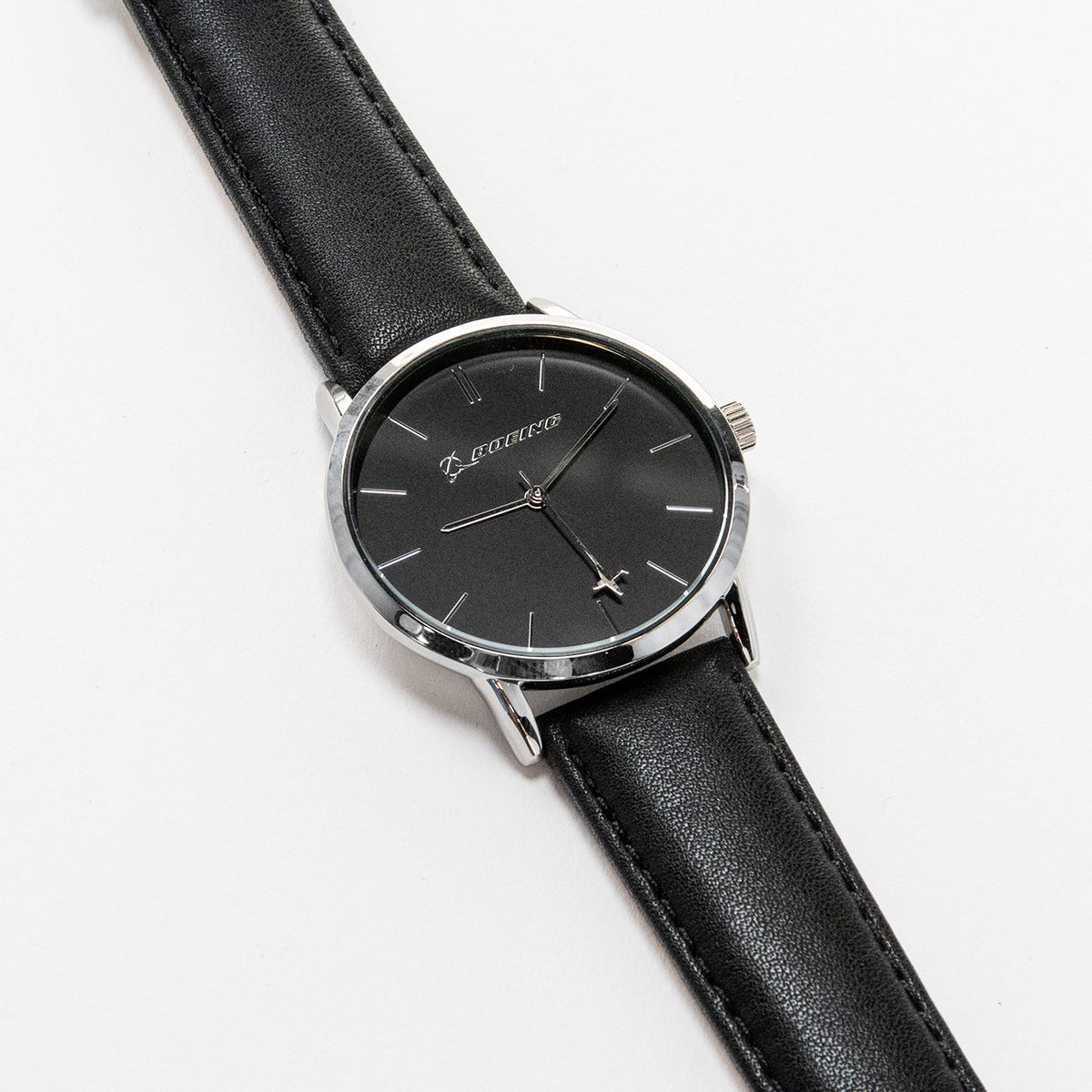 The Boeing Silver Rotating Airplane Watch by Boeing features a minimalist black dial encased in a silver case with a sleek black leather strap. Silver hour markers and hands move elegantly, showcasing timeless style as it rests gracefully on a white surface.