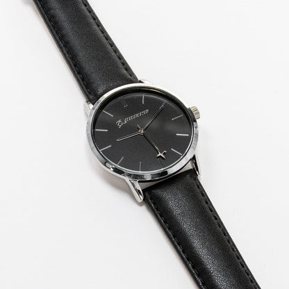 The Boeing Silver Rotating Airplane Watch by Boeing features a minimalist black dial encased in a silver case with a sleek black leather strap. Silver hour markers and hands move elegantly, showcasing timeless style as it rests gracefully on a white surface.