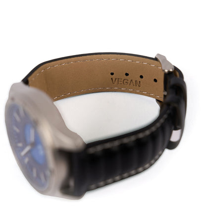 A close-up of The Boeing Store's Boeing Symbol Men's Solar Watch 42 mm shows a black strap with silver buckle, tan vegan AppleSkin lining embossed “VEGAN,” and a blue face powered by precise Japanese solar movement.