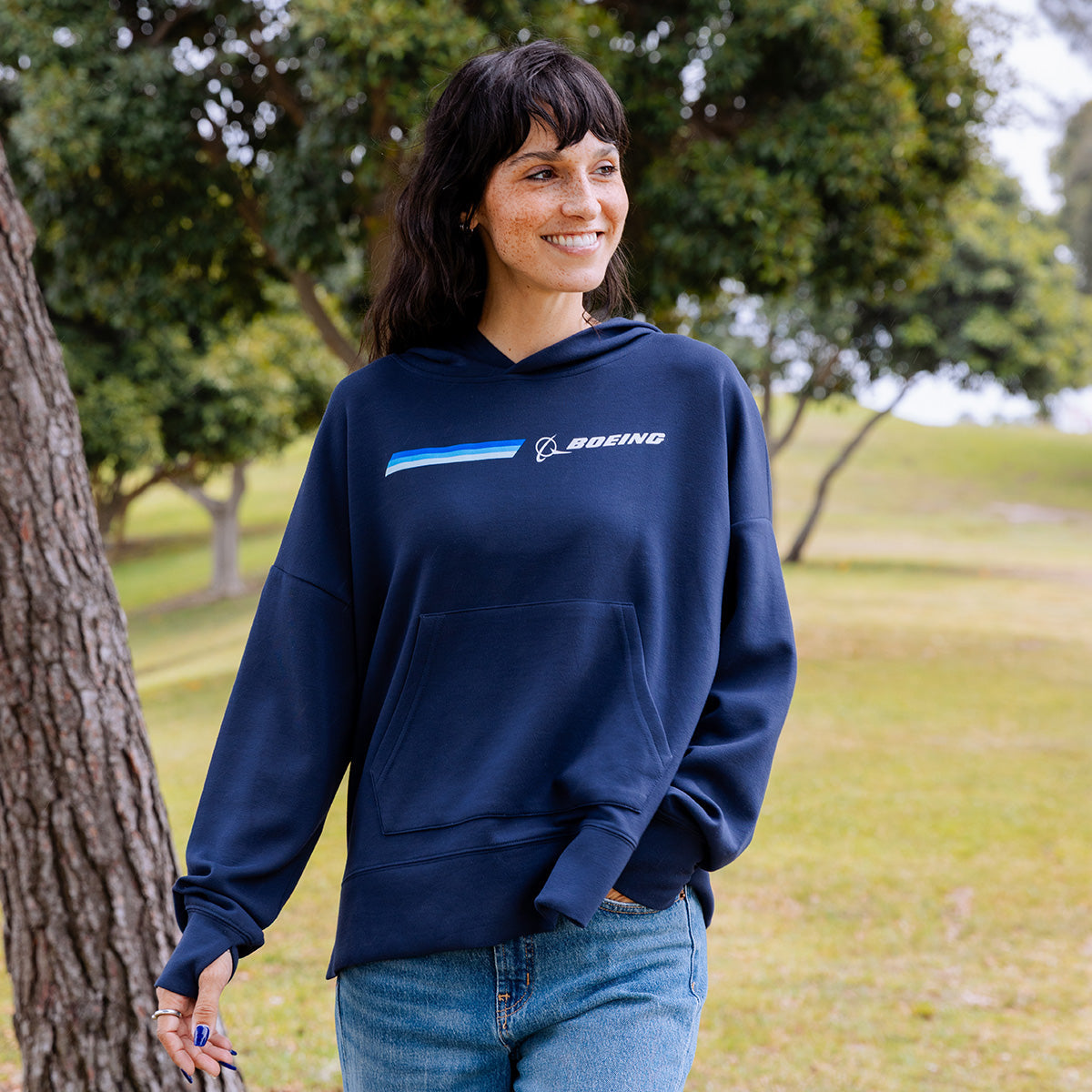 A woman outdoors in a grassy park smiles while wearing the Boeing Women's Signature Logo Stripe Hoodie by BOEING with thumb holes and jeans, highlighting a relaxed, casual look with trees in the background.
