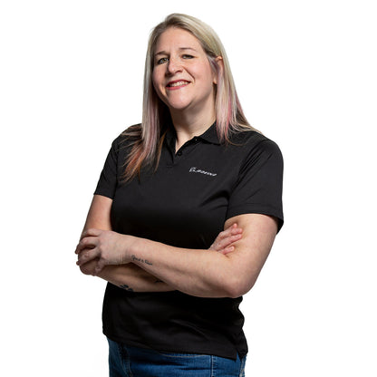 A person with long blonde hair stands confidently, arms crossed, wearing a sleek Boeing Signature Logo Womens Mesh Tech Polo by The Boeing Store and blue jeans. Their poised demeanor is highlighted against a pristine white background.