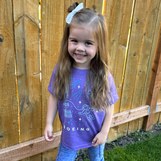 A young girl with long hair stands smiling by a wooden fence, wearing a Boeing Space Doodle Toddler T-Shirt paired with blue pants.