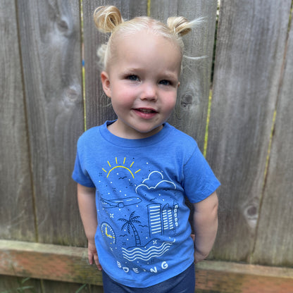 A smiling young child with blond hair styled in buns stands before a wooden fence, wearing a blue Boeing Commercial Doodle Toddler T-Shirt featuring a screen-printed graphic of the sun, clouds, palm trees, and buildings.