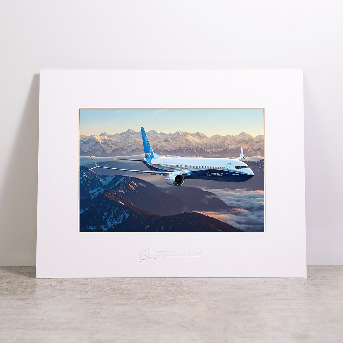 Bring the beauty of aviation to your space with the Boeing 737-10 Matted Print - Small from Boeing, featuring a high-resolution image of a Boeing airplane soaring above snow-covered mountains at sunrise. This professionally matted print includes the official Boeing logo embossed below the image.