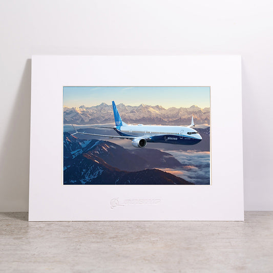 Bring the beauty of aviation to your space with the Boeing 737-10 Matted Print - Small from Boeing, featuring a high-resolution image of a Boeing airplane soaring above snow-covered mountains at sunrise. This professionally matted print includes the official Boeing logo embossed below the image.