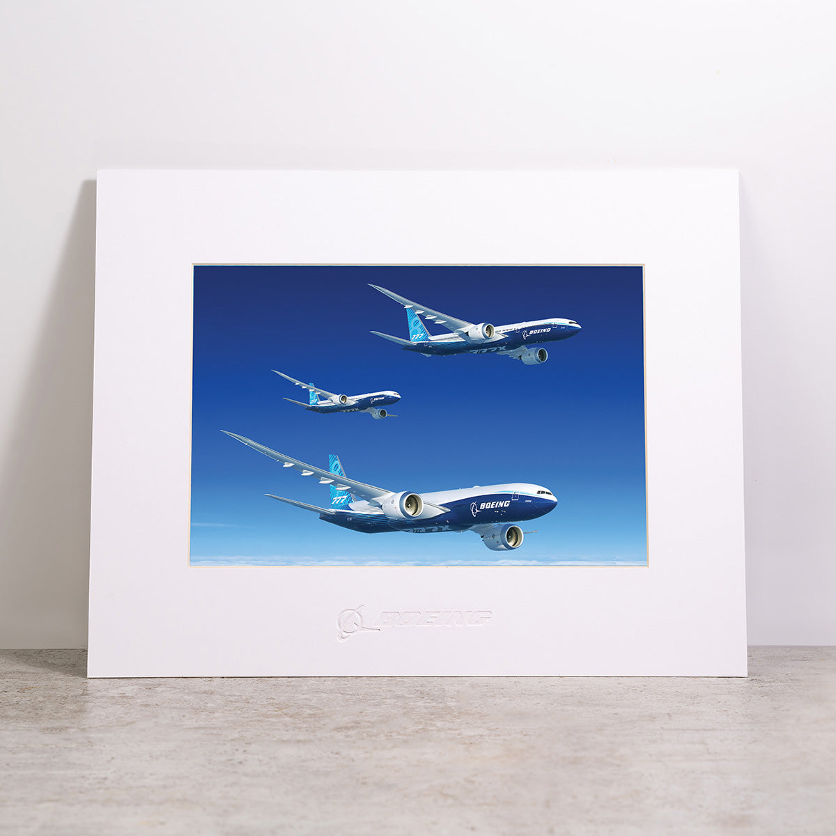 Introducing the Boeing 777X Family Matted Print - Small by Boeing: a high-resolution print featuring three Boeing airplanes in flight at varying angles, set against a clear blue sky. This striking artwork is presented in a white matted frame and features the iconic Boeing logo embossed at the bottom.