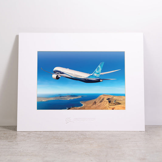 The Boeing 787-9 Matted Print - Large by Boeing showcases a striking photograph of a 787 Dreamliner flying over a rugged coastline and deep blue sea beneath a clear sky. The white matting is elegantly embossed with the Boeing logo, and the high-quality digital print ensures an impressive presentation.