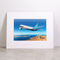 The Boeing 787-9 Matted Print - Large by Boeing showcases a striking photograph of a 787 Dreamliner flying over a rugged coastline and deep blue sea beneath a clear sky. The white matting is elegantly embossed with the Boeing logo, and the high-quality digital print ensures an impressive presentation.