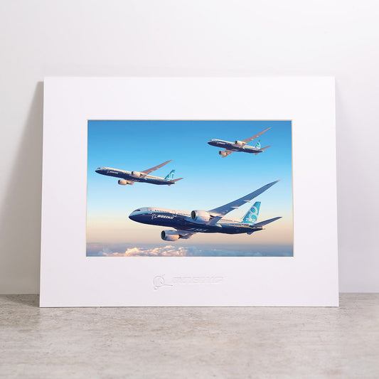 The Boeing 787 Dreamliner Family Matted Print - Small by Boeing showcases a high-resolution image of three Boeing airplanes soaring above the clouds in a vivid blue sky. Printed on premium paper and elegantly matted, this print features an embossed Boeing logo at the bottom for a refined touch.