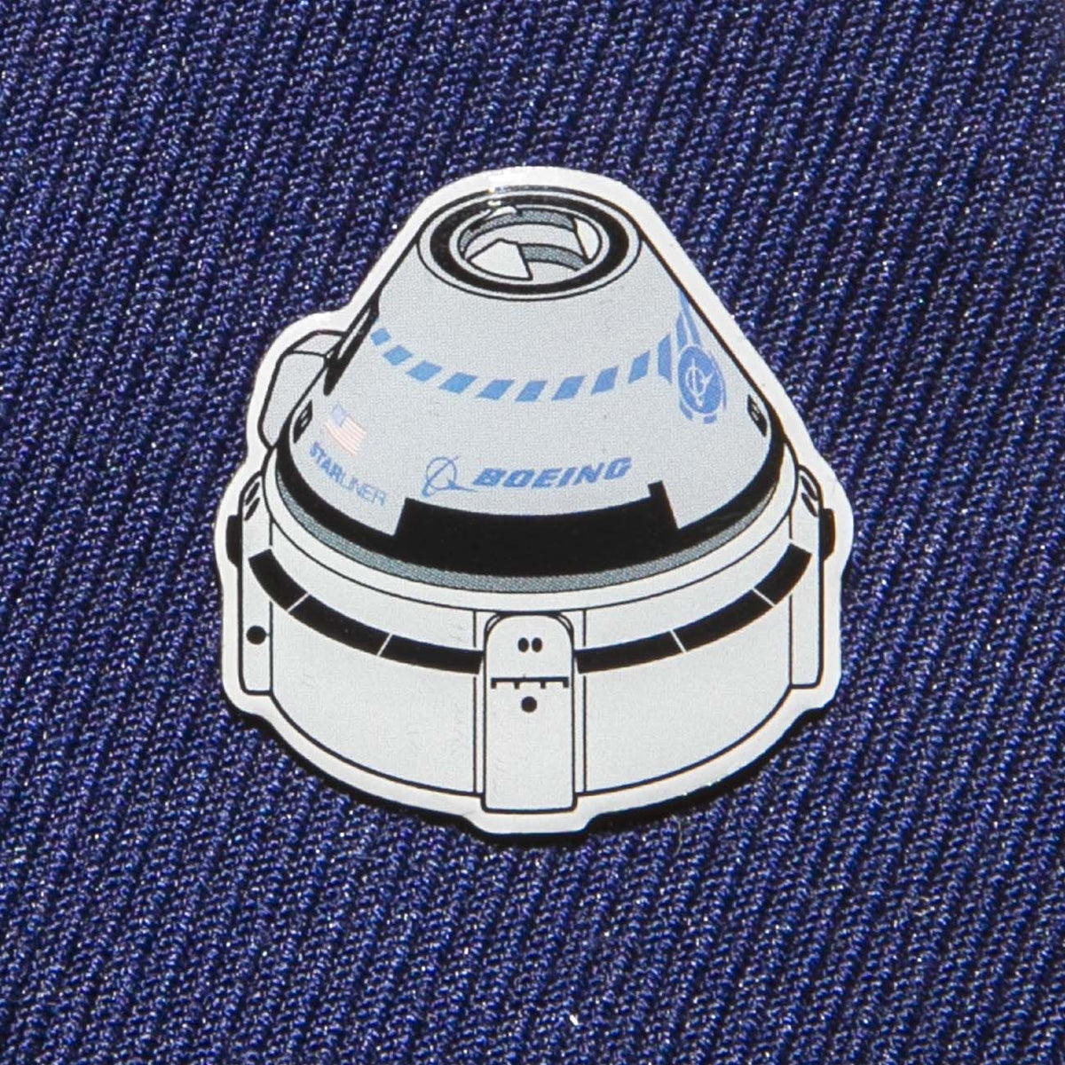 A Boeing Illustrated Starliner Lapel Pin by The Boeing Store features a spacecraft design on blue fabric. It showcases an epoxy finish and small logos, including the Boeing logo, for a realistic touch.