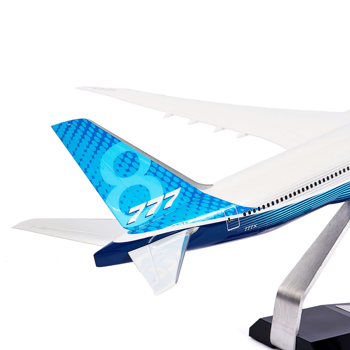 The Boeing Store offers a finely crafted 1:144 Boeing Unified 777-8 model. Its sleek, white wings contrast with the stylish blue-patterned tail fin featuring a stylized 777. Elegantly mounted on a metal stand, this twin-engine jet model is perfect for aviation enthusiasts.