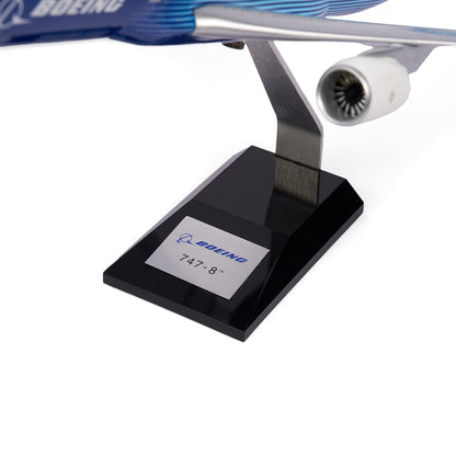 A close-up of the Boeing Unified 747-8i 1:144 Model by The Boeing Store on a black stand shows its blue livery, detailed engine, and fuselage with the Boeing logo—celebrating this premium jet for high-volume routes.