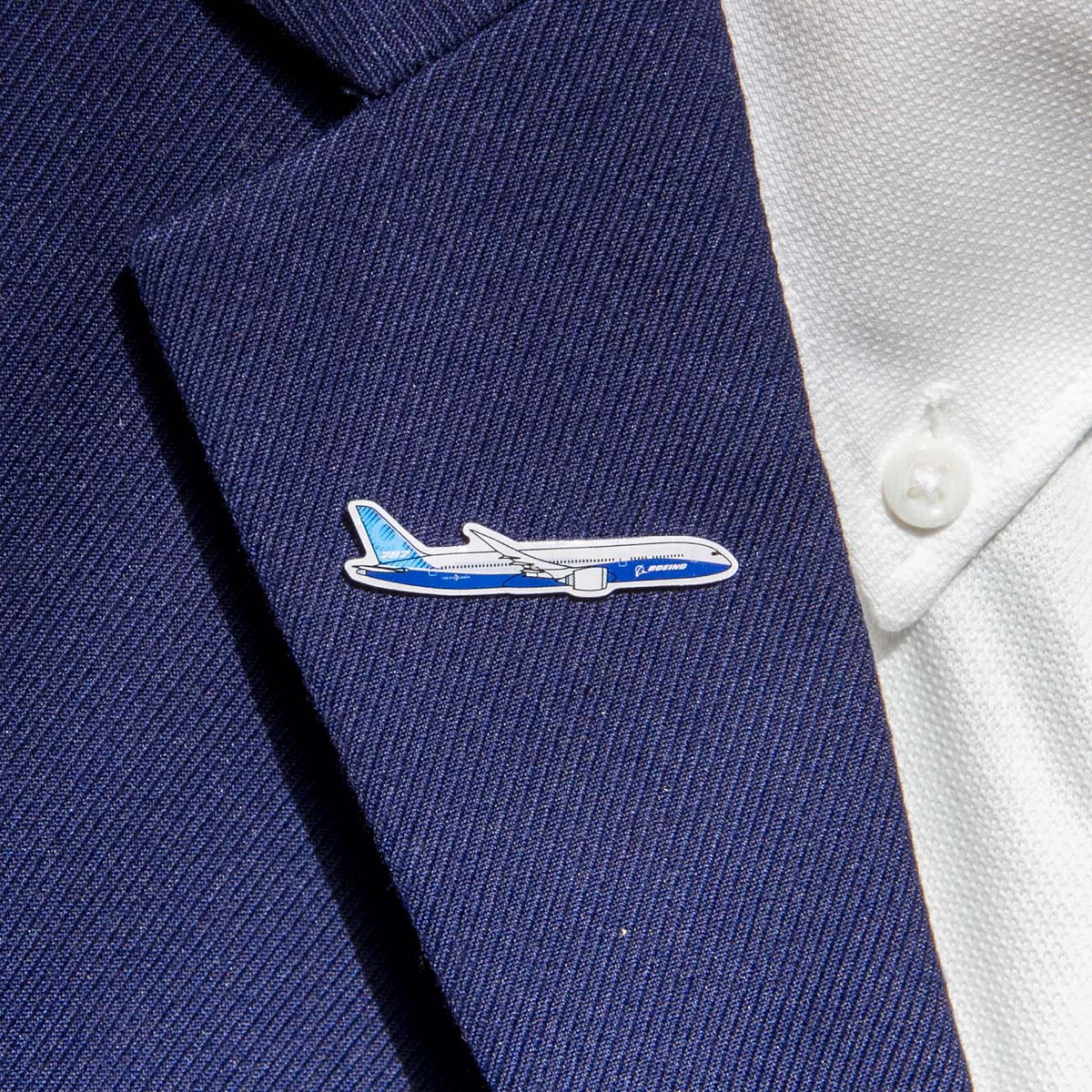 A close-up of a navy blue suit jacket paired with a white shirt highlights the Boeing Illustrated 787 Lapel Pin from The Boeing Store, featuring a silver-tone design of a blue and white airplane that exudes elegance and style.