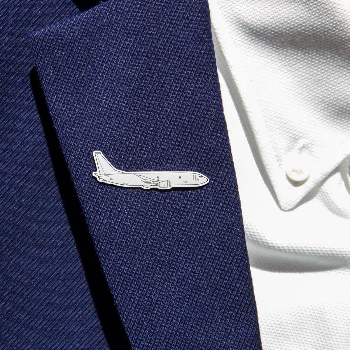 A person wears a dark blue suit jacket featuring the Boeing Illustrated P-8 Lapel Pin by The Boeing Store, shaped like an airplane and resembling the Boeing logo, on the lapel. A white textured shirt completes the look.