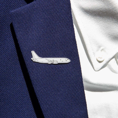 A person wears a dark blue suit jacket featuring the Boeing Illustrated P-8 Lapel Pin by The Boeing Store, shaped like an airplane and resembling the Boeing logo, on the lapel. A white textured shirt completes the look.