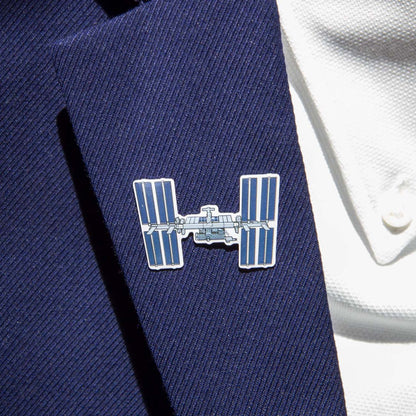 A person wearing a blue pinstripe suit jacket and white shirt showcases The Boeing Stores Boeing Illustrated ISS Lapel Pin. The silver-tone pin, featuring enamel details and an epoxy finish, adds sophistication to any outfit.