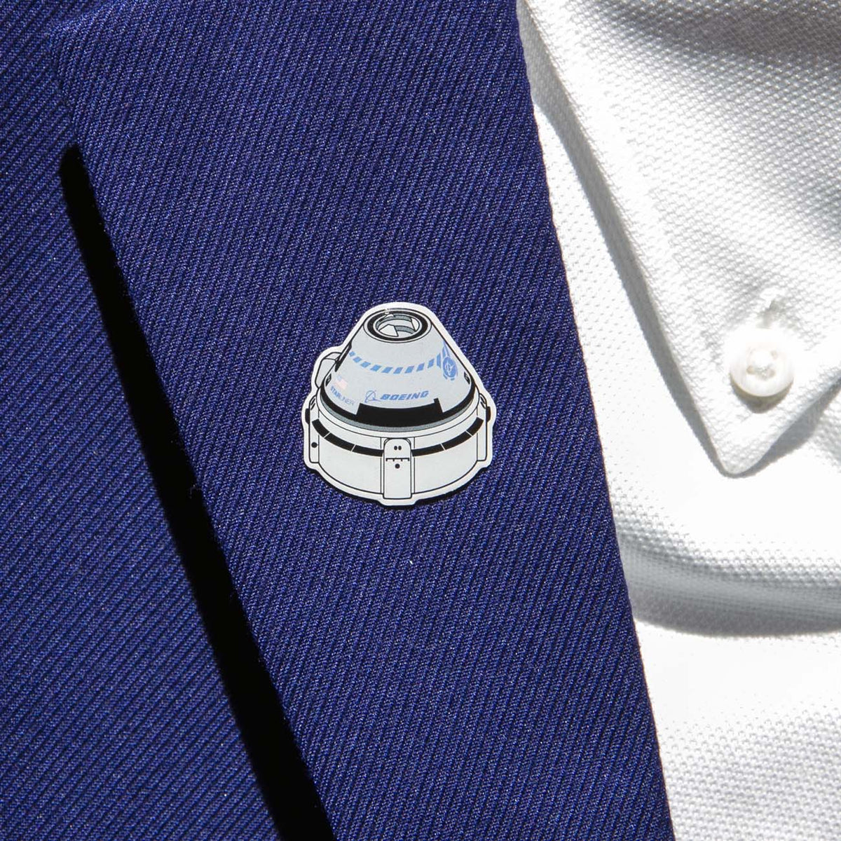 A Boeing Illustrated Starliner Lapel Pin with an epoxy finish and a small logo from The Boeing Store is attached to a blue suit jacket worn over a white textured shirt with a button showing.