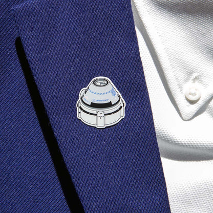 A Boeing Illustrated Starliner Lapel Pin with an epoxy finish and a small logo from The Boeing Store is attached to a blue suit jacket worn over a white textured shirt with a button showing.