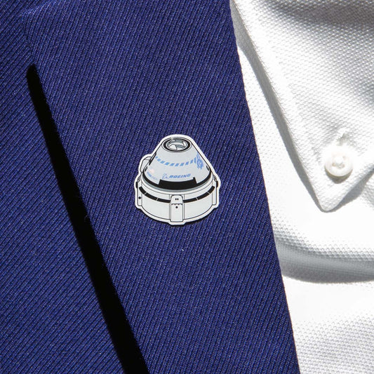 A Boeing Illustrated Starliner Lapel Pin with an epoxy finish and a small logo from The Boeing Store is attached to a blue suit jacket worn over a white textured shirt with a button showing.