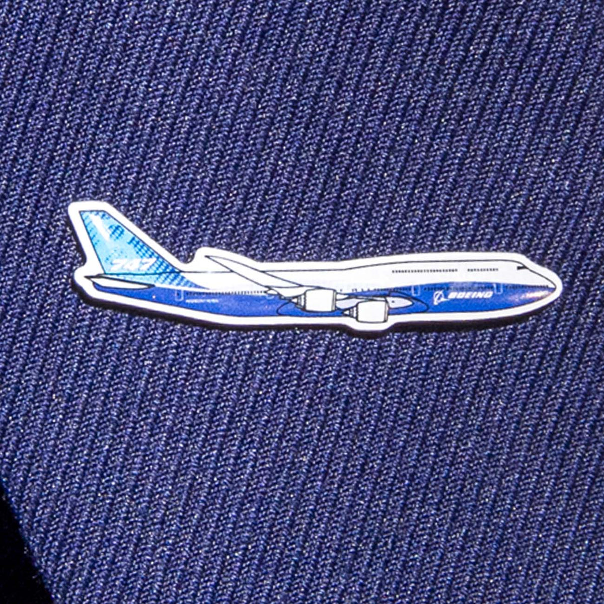 The Boeing Stores Boeing Illustrated 747 Lapel Pin is silver-toned and shaped like a blue and white airplane, featuring an elegant epoxy finish on a dark blue textile background.