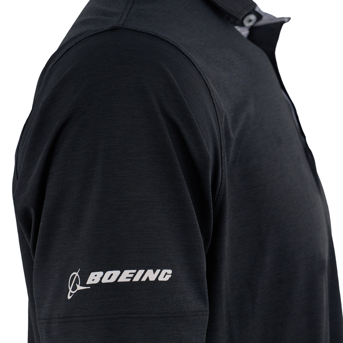 A close-up of someone wearing the Boeing T-7A Red Hawk Series Men’s Polo by BOEING, featuring a black design, white logo and name on the left sleeve, buttoned placket, moisture-wicking fabric, and a plain white background.