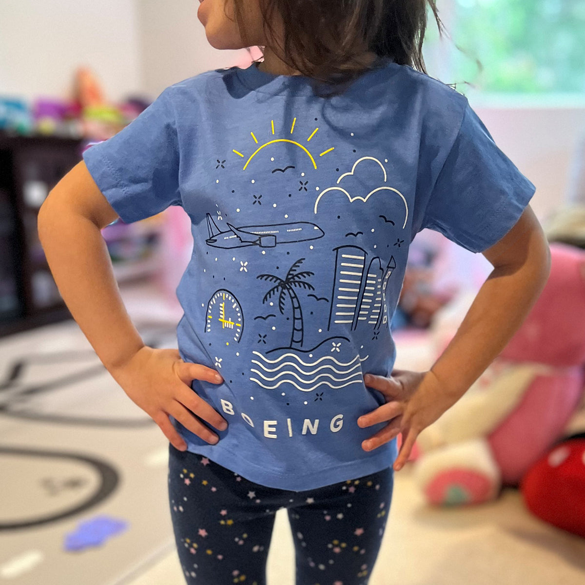 A confident child stands with hands on hips, wearing a Boeing Commercial Doodle Toddler T-Shirt featuring a screen-printed design of sun, clouds, plane, palm trees, and waves. In the vibrant playroom background are soft toys and colorful décor.
