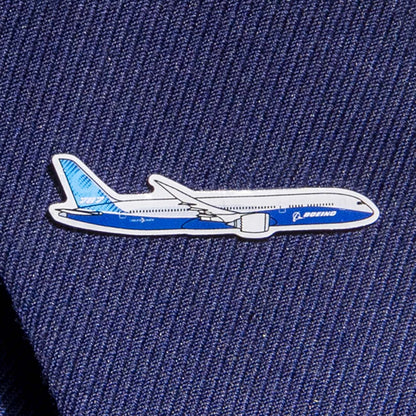 A silver-toned Boeing Illustrated 787 Lapel Pin from The Boeing Store, shaped like a Boeing airplane with an epoxy finish, adorns dark blue fabric. Its enhanced blue and white colors feature the Boeing logo prominently on the side.