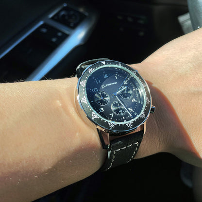 A wrist sports a Boeing Black Leather Watch: round face, silver markings, iconic Boeing logo. It features a stainless steel case and black leather strap with white stitching against a car interior backdrop. Available at The Boeing Store.
